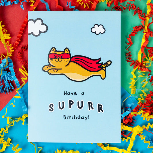 The Have A Supurr Birthday Cat Card on a red, blue and green background with red, yellow and blue crinkle card confetti. The pastel blue a6 birthday card of a smiling orange cat flying through a cloudy sky wearing a superhero cape and mask, underneath in black text reads 'have a supurr birthday!'.