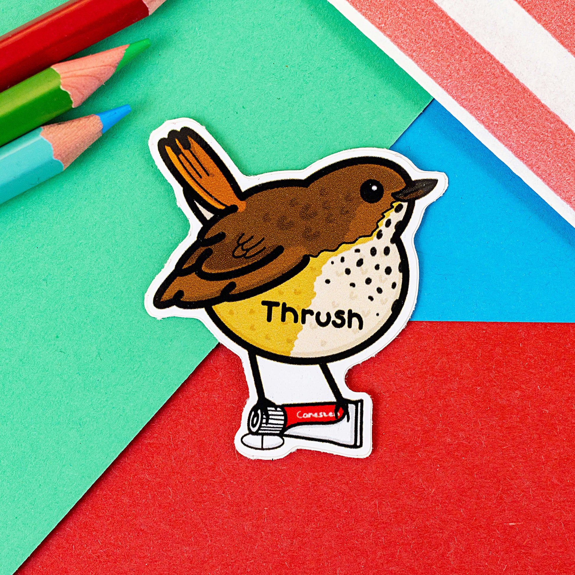 The Thrush Bird Sticker on a red, blue and green background with colouring pencils and red stripe candy bag. The brown thrush bird is stood smiling on a tube of thrush canesten cream and tablet, across its middle in black text reads 'thrush'. The hand drawn design is raising awareness for thrush.