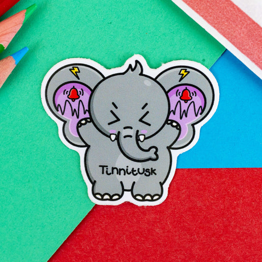 The Tinnitusk Elephant Sticker - Tinnitus on a red, blue and green background with colouring pencils and red stripe candy bag. A grey elephant shaped vinyl sticker with big ears with purple on the inside of them and black squiggly lines with red ringing alarm bells above the lines and yellow lightening bolts above the bells. The elephant has its eyes screwed shut and its arms up. 'Tinnitusk' is written in black across its middle.