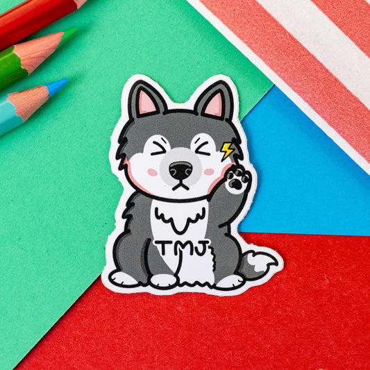 The Tempawromandibular Disorder Dog Sticker - Temporomandibular Disorder TMJ on red, blue and green card with colouring pencils and red stripe candy bag. The grey husky dog shaped vinyl sticker has a pained expression holding up its paw to its jaw with a yellow lightning bolt, across its middle in black reads 'TMJ'. The hand drawn design is raising awareness for Temporomandibular Disorder TMJ