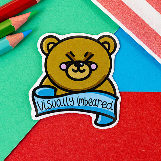 Visually impaired sticker on a red and blue background. The sticker has an adorable brown teddy bear with rosy cheeks. The bear's eyes are closed and there is a blue banner under the bear with 'Visually imbeared' written across it in black writing.