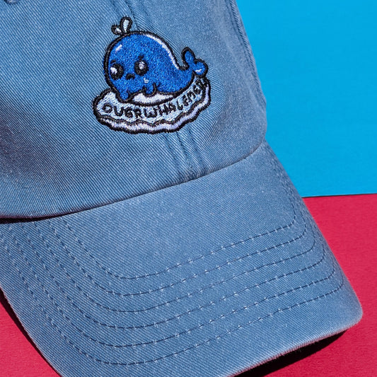Close-up of Overwhalemed cap by Innabox on bright background. Features a teary blue whale embroidered patch and soft, vintage light blue denim.