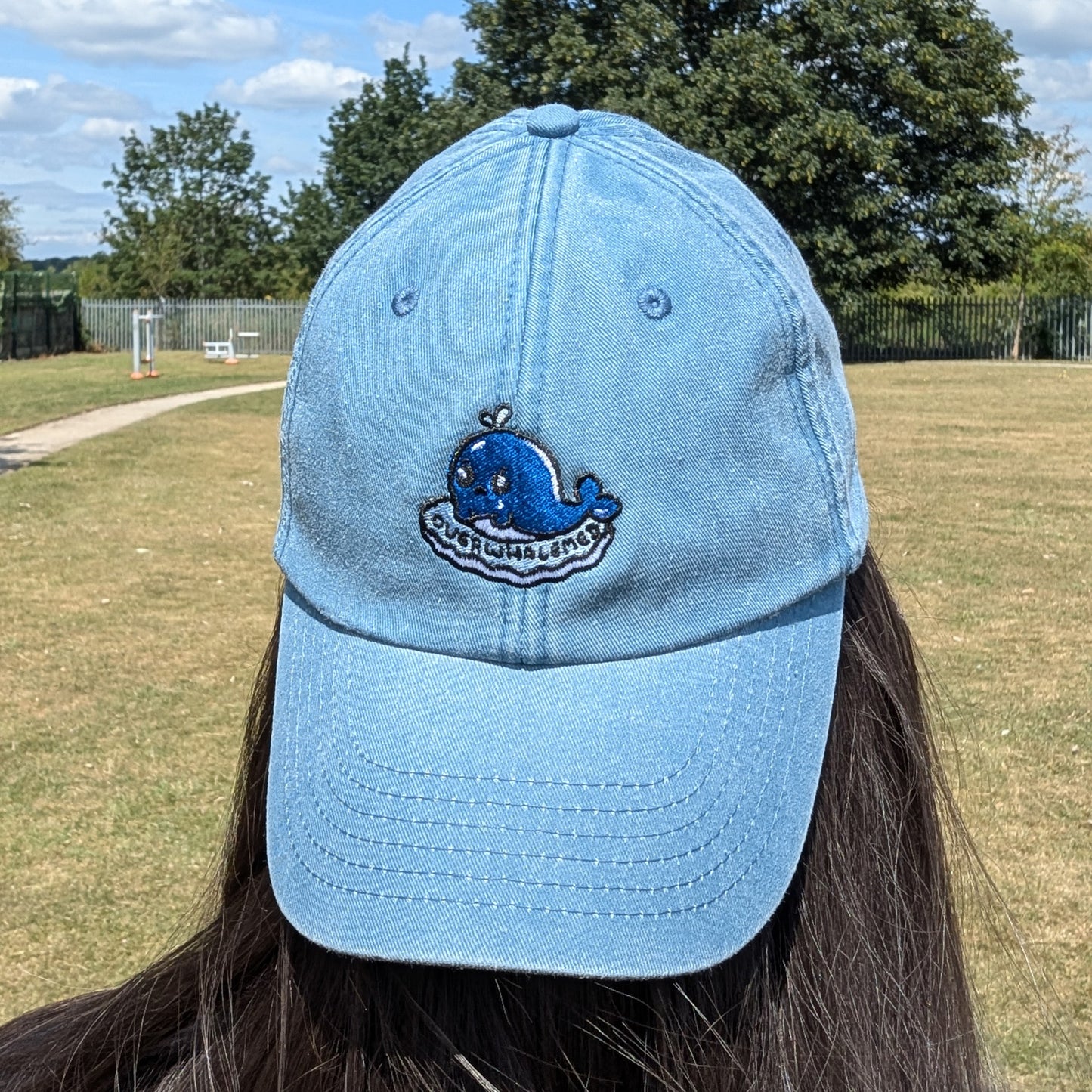 Back view of a light blue washed denim Overwhalemed cap worn outdoors, showing the adjustable fabric strap with brass-effect buckle. Designed by Innabox.