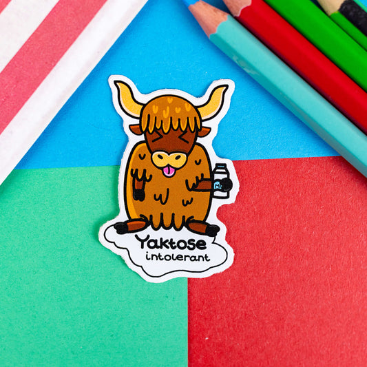 A sticker with a Yak and drinking milk looking disgusted with the text Yaktose Intolerant the background is green, blue, and red with coloured pencils at the side of frame and a red and white striped bag in the other corner