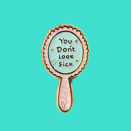 You Don't Look Sick Mirror Enamel Pin on a blue background. The hand mirror shaped baby pink glittery enamel pin has gold text on it that reads 'you don't look sick'. The hand drawn design is made to raise awareness for invisible illnesses  