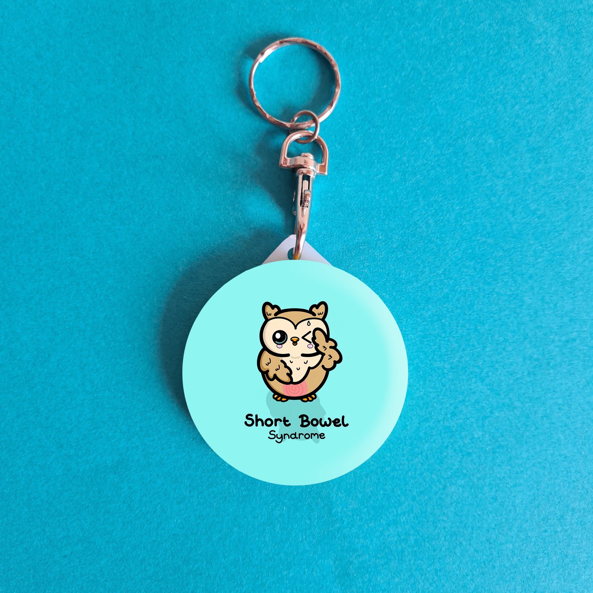 a light blue circle shaped keyring with a little cute brown owl with a red tummy and a sweat droplet coming from it's forehead with one eye closed. 'Short Bowel Syndrome' is written underneath the owl. The background of the photo is blue