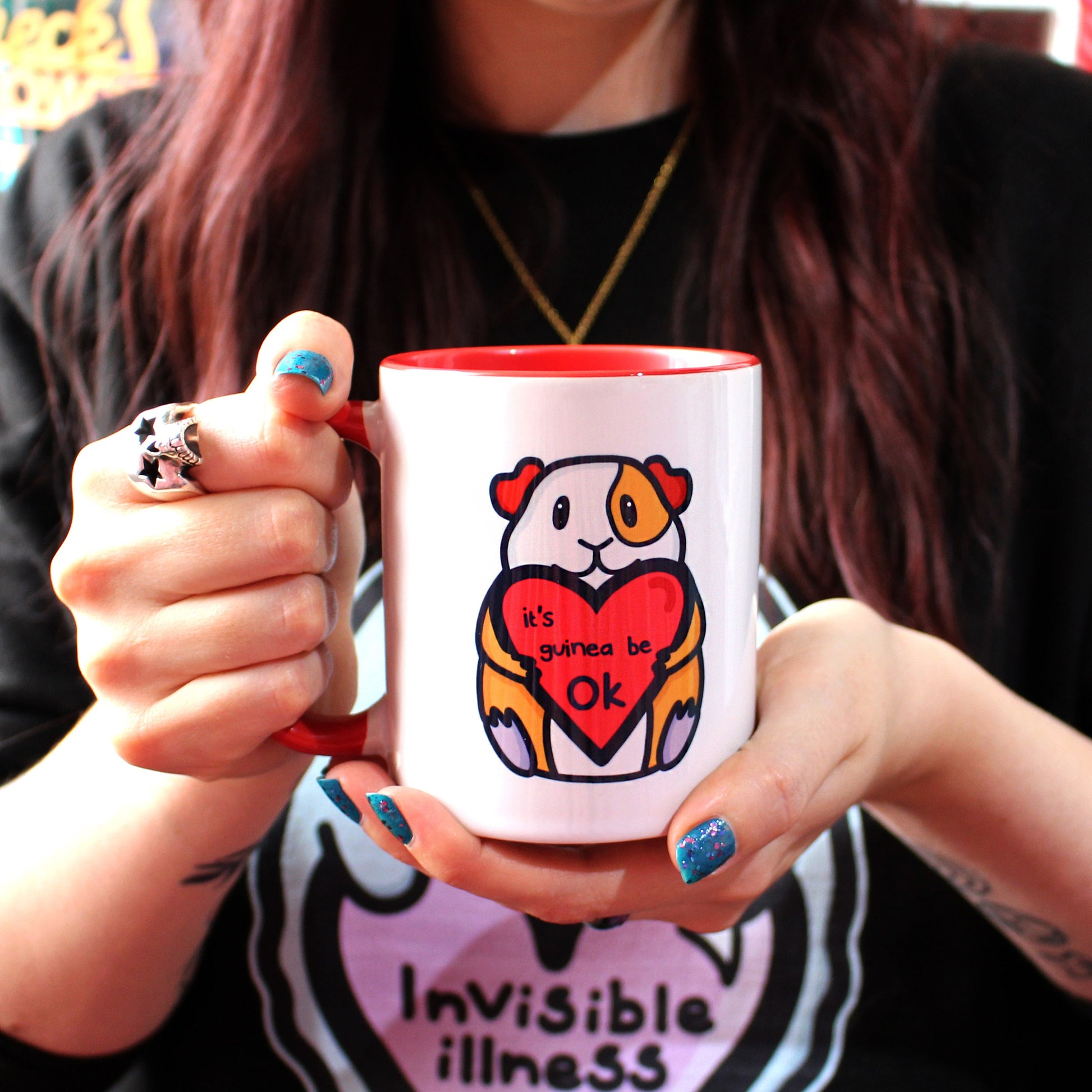 The It's Gonna Be OK Guinea Pig Mug being held up by Nikky, the owner of Innabox, who has brown long hair, blue nail varnish and large silver skull ring. The white mug has a red inside and handle with a front print of a orange and white guinea pig smiling holding a red heart with black text reading 'it's guinea be ok'. The hand drawn design is a reminder of staying positive.