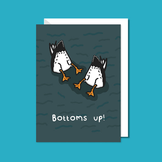 A card with a dark murky sea base with two black and white seagull bottoms up in the air in the sea with the text 'bottoms up!' underneath them. The celebration card is on a blue background with a white envelope underneath. 