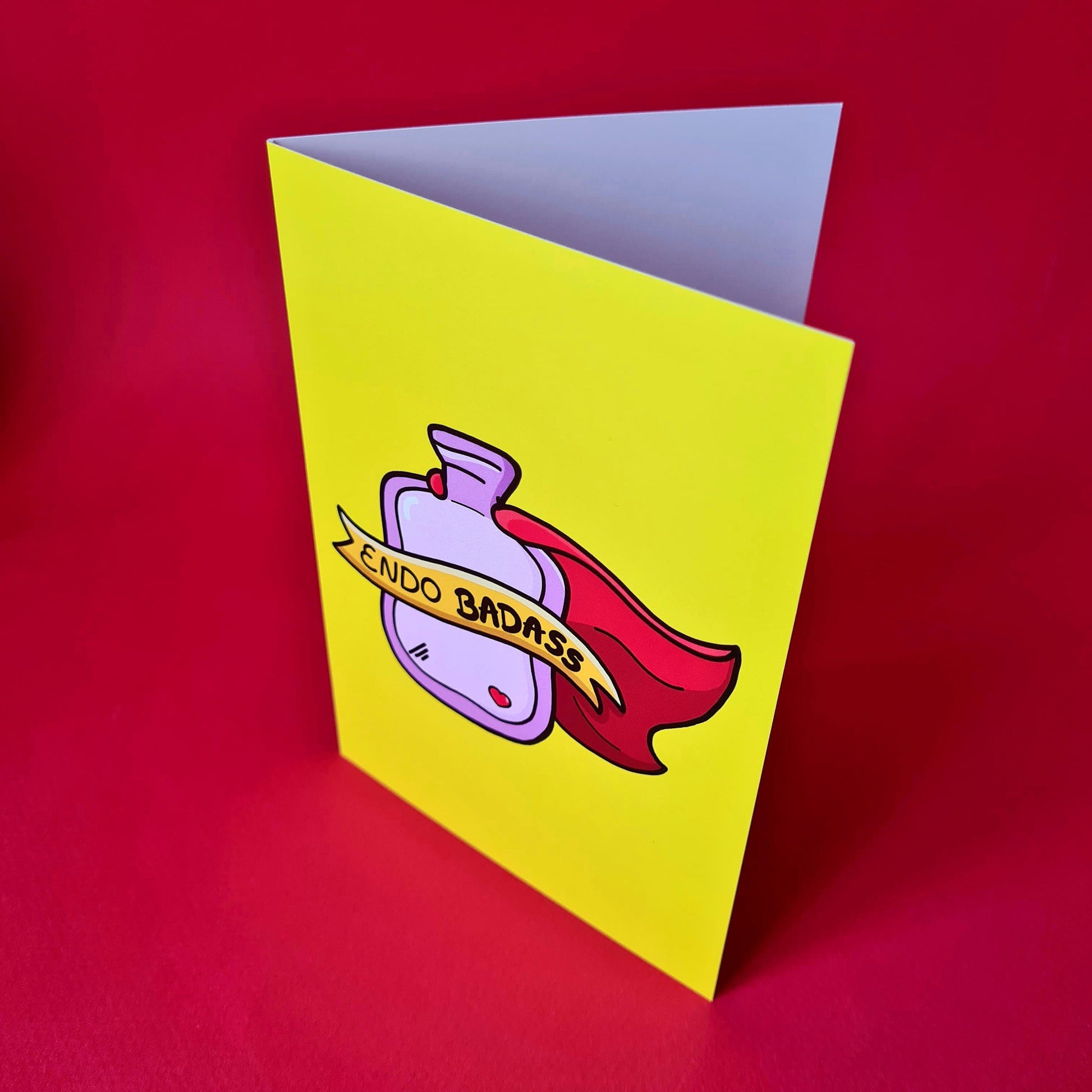The Endo Badass Card - Endometriosis on a red background. The yellow based card features a pink hot waterbottle with a small red heart, a red cape and a yellow banner across reading 'endo badass'. Hand drawn design is raising awareness for Endometriosis and pelvic pain.